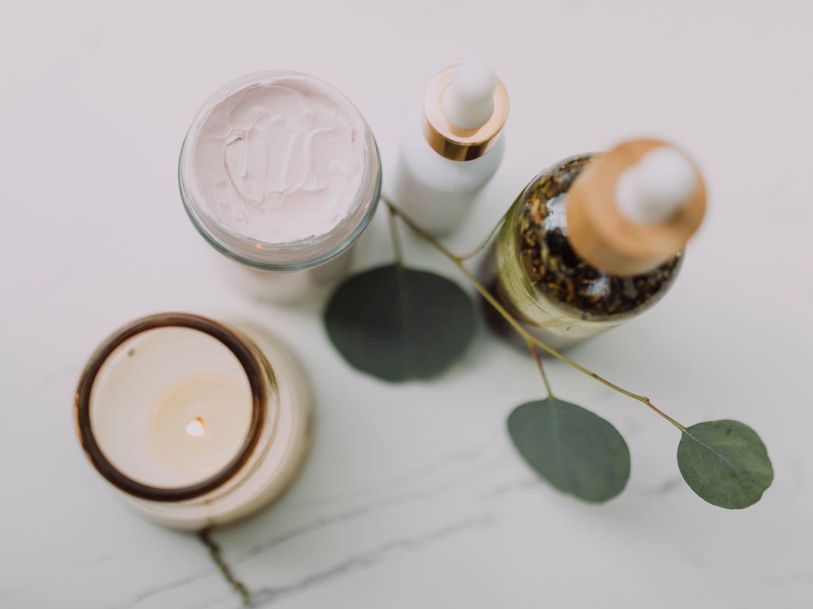A flat lay of skincare products, a cream jar, serum bottles, a candle and eucalyptus leaves on marble